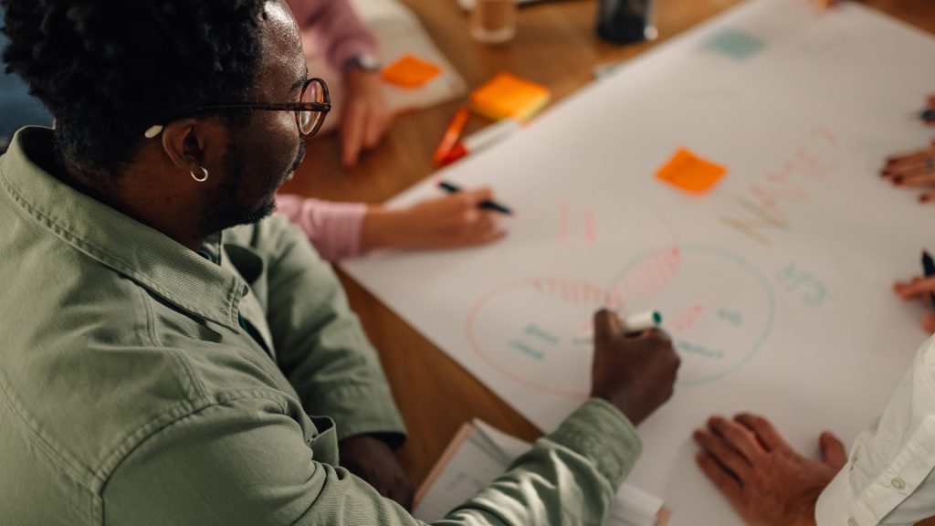Person seen contributing to brainstorming session with team. They are writing on large piece of paper using ink pens.