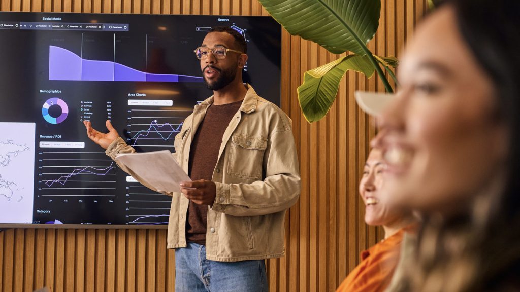 Person presenting to team in meeting room. On screen there are graphs and data.