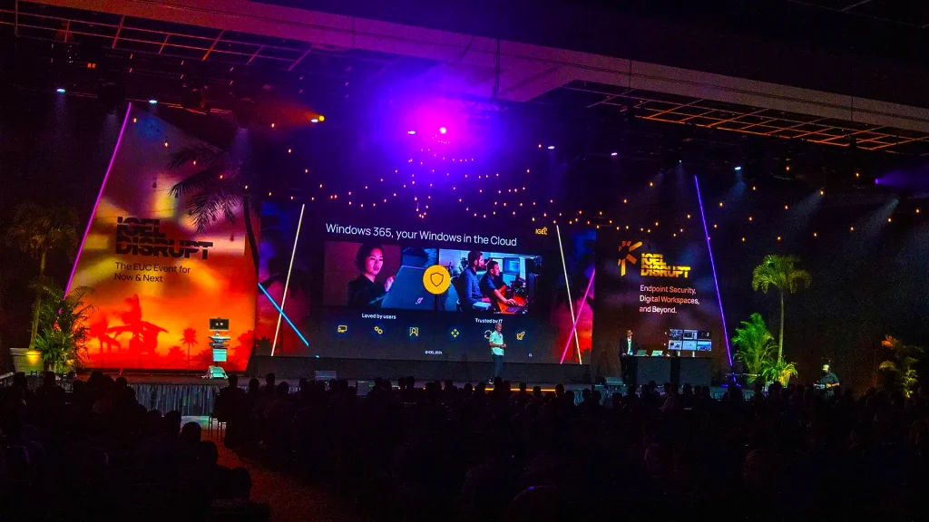Disrupt stage with colourful lighting, palm trees and branding on screen.
