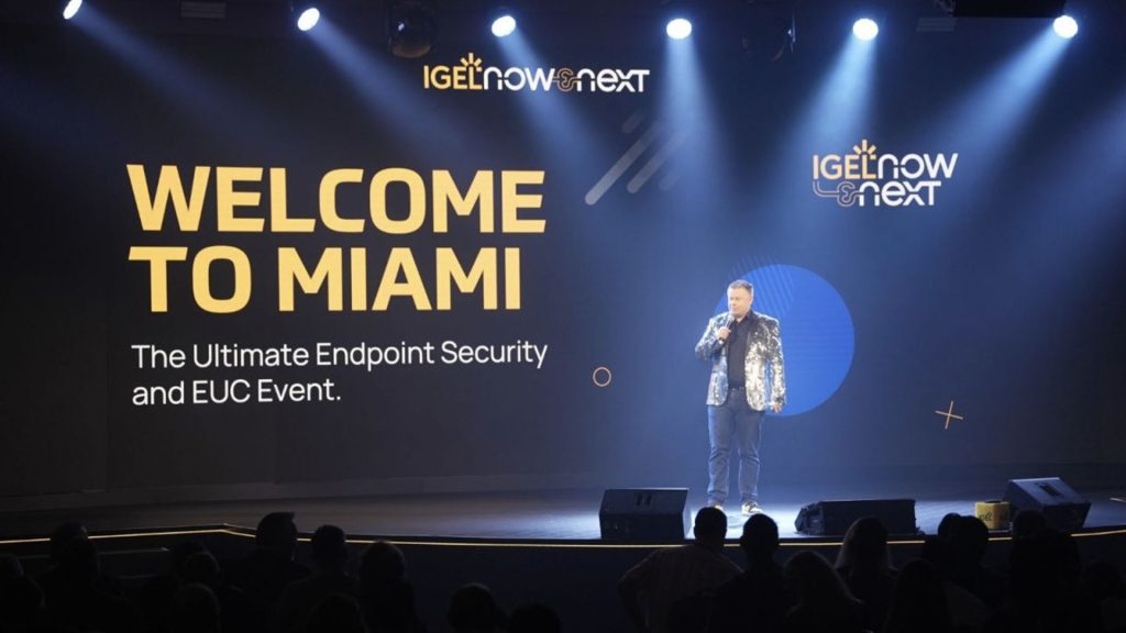 Presenter stands in front of 'Welcome to Miami' screen on stage.